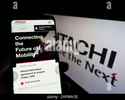 Person holding cellphone with logo of Japanese company K.K. Hitachi Seisakusho on screen in front of business webpage. Focus on phone display. Stock Photo
