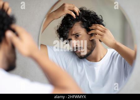 Haircare treatment. Worried indian guy looking at flaky scalp in mirror, examining gray hair and hairloss issue Stock Photo