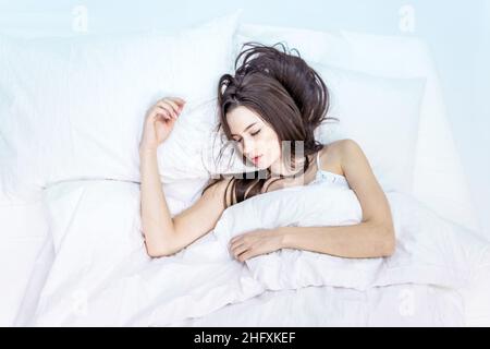 Good morning. Get enough sleep. Bedroom. The brunette sleeps on a white bed. Seeing pleasant dreams. A cute girl in pajamas lies on the bed with her e Stock Photo