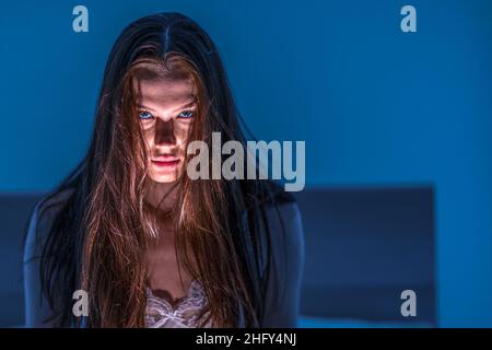 The woman shines a flashlight at herself from below and creates a scary face. Horror. Scare. Scary girl with dark hair Stock Photo