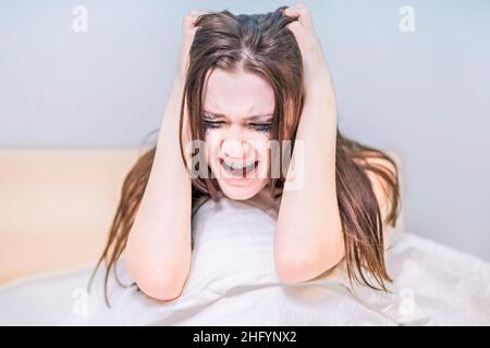 A woman with a face dirty from mascara sits in bed and cries and screams. Get the bad news. Get upset. To suffer. Depression. Heartbroken. Broken hear Stock Photo