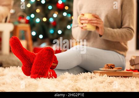 Woman in warm socks with cup sitting on floor at home on Christmas eve Stock Photo