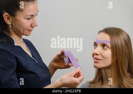 female cosmetician doing the cosmetic kinesio taping of the face. Purple stripes on forehead against wrinkles and aging Stock Photo