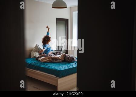 Curly woman in pajamas sitting on bed in front of laptop and watching online lesson while making notes in copybook. Internet technology. Stock Photo