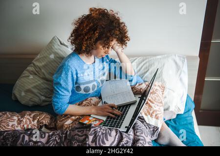 Curly brunette woman sitting in bed making notes in notepad using laptop. Bedtime. Stock Photo