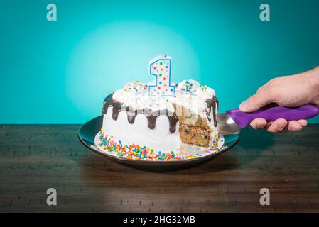 A birthday cake, missing a slice, bears a candle in the shape of the number 1 while a hand cuts another slice. Stock Photo