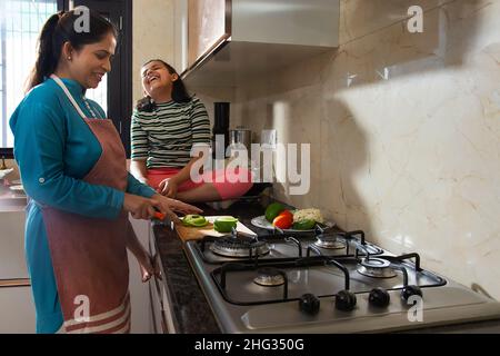 Middle class Indian woman chopping vegetables and her daughter sitting  beside her in kitchen Stock Photo