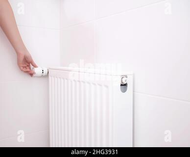 a woman regulates the temperature of the radiator heating with a thermostat. Wall mounted heat radiator in the bathroom Stock Photo