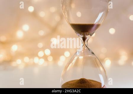 Hourglass on a festive background of blurry garlands and bokeh in creamy beige tones. Christmas, new year, copy space. Waiting for the feast. Stock Photo