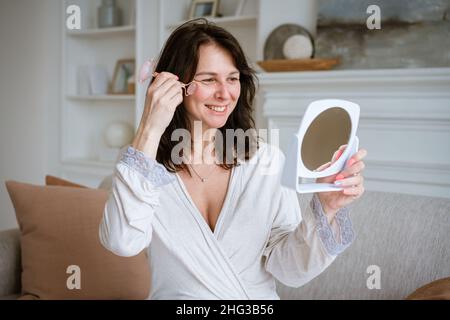Woman using face roller. Sculpting, lifting and anti-aging care. Skin care and beauty at home. Woman using calming quartz roller massager while sitting on sofa in living room looking in mirror Stock Photo