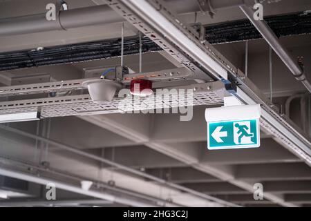 front view closeup of lighted green exit sign hanging on building pipes and pointing the way out of an industrial building Stock Photo