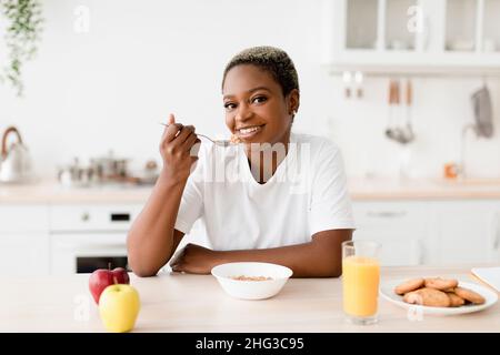 Smiling young attractive black woman eating porridge sits at table with cookies, juice and apples Stock Photo