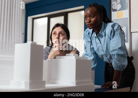 Project engineers women colleagues looking inspired in front of 3d residential maquette in architectural modern office. Focused professional architects team inspecting skyscraper white foam model. Stock Photo