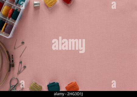 Embroidery background. Cross-stitch accessories are lying on a pink canvas, top view. Stock Photo