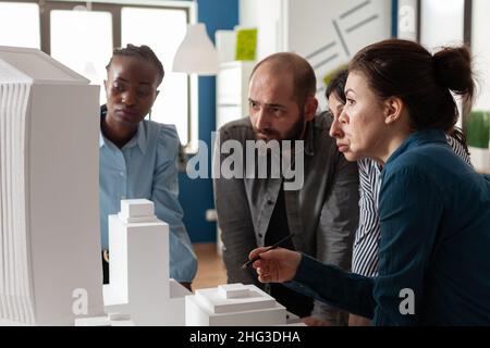 Professional architects discussing architecture design of skyscraper foam scale maquette in architectural office. Team of engineers working together on group project at desk with buildings model. Stock Photo