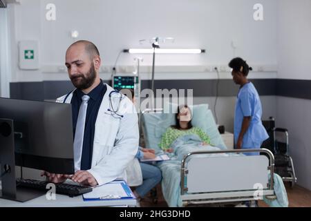 Medical doctor using personal computer to complete patient admission chart while nurse and physician talk with patient in hospital ward. Medic with stethoscope looking at lab results on screen. Stock Photo