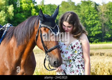 Portrait of a smiling caucasian teenage girl in a dress, holding a bay horse with snaffle, saddle and black ear bonnet. Stock Photo