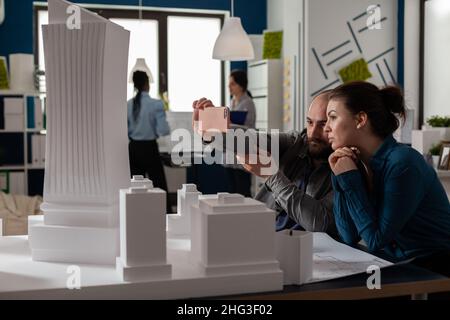 Team of two architect colleagues using smartphone in videocall conference with client presenting desk with buildings models. Architectural engineers working together with smart phone and maquette. Stock Photo