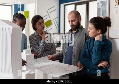 Team of engineers looking at architect holding blueprints for construction project in modern architectural office. Professional engineers analyzing paper plans for building in front of foam maquette. Stock Photo