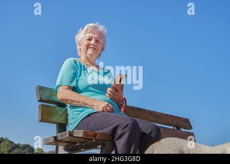 Low angle of optimistic old female sitting on bench near dog and using cellphone under cloudless blue sky, in sunny day and looking at camera Stock Photo