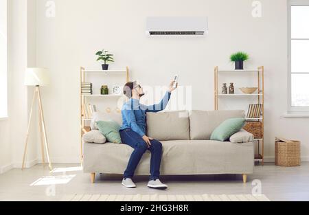 Young man turns on air conditioner with remote control while relaxing on couch at home. Stock Photo