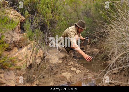 Young caucasian male adventurer collecting water from small pond amidst rocks and plants in forest Stock Photo