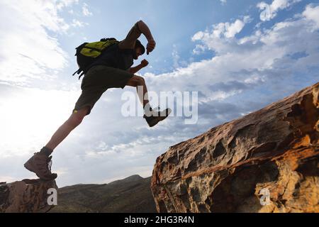 Low angle view of athletic young male caucasian hiker jumping on rocky cliffs during sunny day Stock Photo