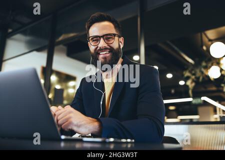 Successful entrepreneur smiling at the camera in an office. Happy young businessman listening to music while working on a laptop. Cheerful businessman Stock Photo