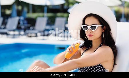young woman with straw hat and sunglasses spraying sunscreen on arm near swimming pool Stock Photo
