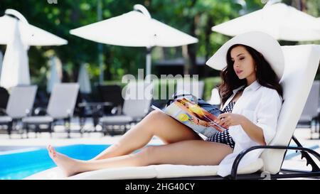 brunette woman in straw hat reading magazine while resting on lounger near swimming pool Stock Photo