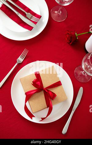 Valentine's day dining table concept. Plate with gift box, utensil, bottle of wine on red background. Flat lay, top view. Stock Photo