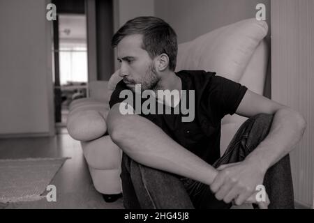 Young pensive bearded man sits on floor near sofa in t-shirt and jeans. Stock Photo