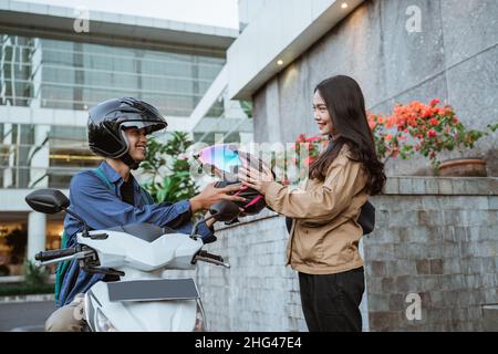 Asian woman receiving helmet from man riding motorbike on roadside Stock Photo