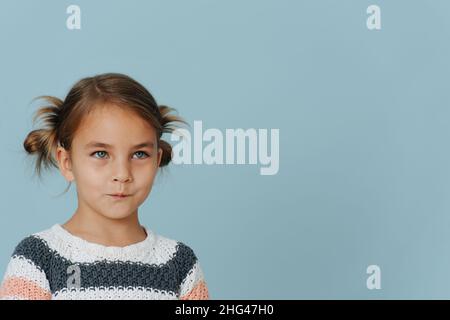 Resentful tight-lipped little girl in striped sweater, hair in buns over blue background. Looking at someone. Off-centered, copy space. Stock Photo