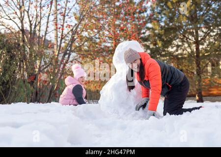 Portrait of small girl and mother crawling on snow near snowman on backyard in evening with rowan and fir trees in background. Parents spending time Stock Photo