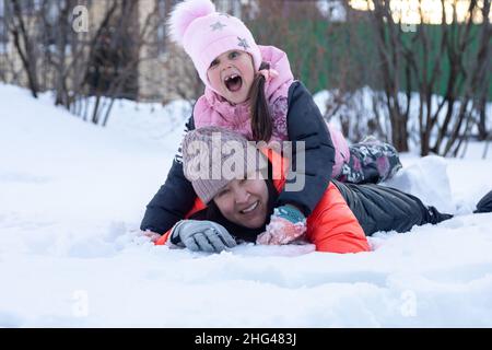 Middle aged mother with little daughter lying on snow together smiling with teeth in park in evening with trees in background. Parents spending time Stock Photo