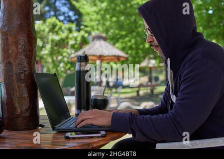 latin man wearing a hoodie working on his laptop and drinking mate outdoors while he is on vacation. Concept remote work and travel . Horizontal Stock Photo