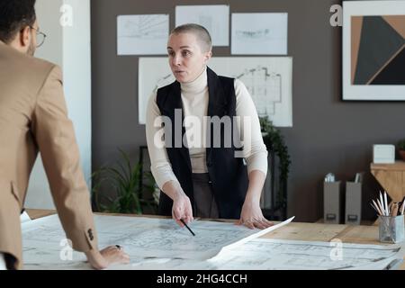 Confident mature female architect talking to male colleague while pointing at sketch on blueprint at working meeting Stock Photo