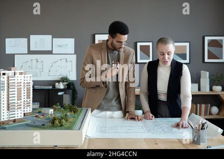 Two elegant architects or engineers discussing sketch on blueprint while standing by large table with house layout Stock Photo