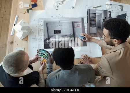 Overview of contemporary designers bending over workplace with blueprint and photos of interior while discussing color palette Stock Photo