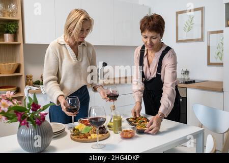 Two mature women swerving appetizers and glasses with wine on dining table in the kitchen Stock Photo