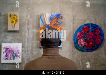 Rear view of young African man looking at collection of abstract paintings on wall while visiting art exhibition Stock Photo