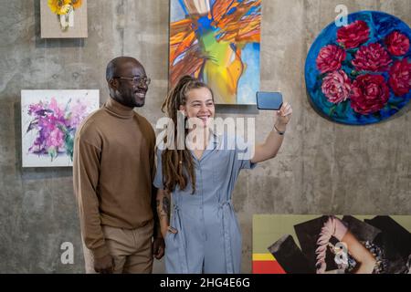 Happy young intercultural couple making selfie in art gallery against wall with collection of paintings Stock Photo