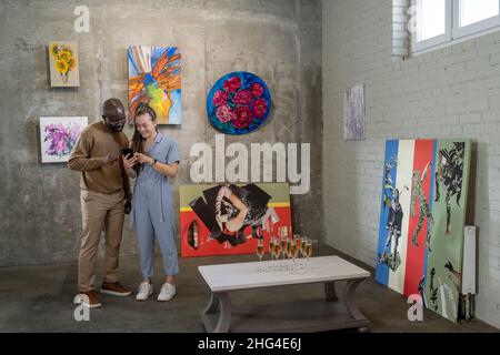 Young intercultural couple looking through their photos in smartphone while standing by wall with collection of paintings Stock Photo