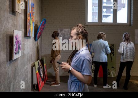 Side view of young female with dreadlocks looking at painting on wall against other guests of modern art gallery Stock Photo
