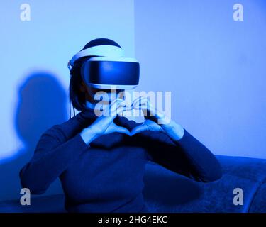 Beautiful woman using vr headset shows heart shape with hands at home in blue light. The concept of virtual relationships. Virtual love concept. Stock Photo