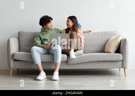 Happy young Asian spouses shopping online via laptop, using credit card, sitting on sofa at home, empty space Stock Photo