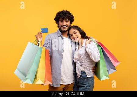 Loving indian couple posing with credit card and colorful shopping bags, making purchases together, yellow background Stock Photo