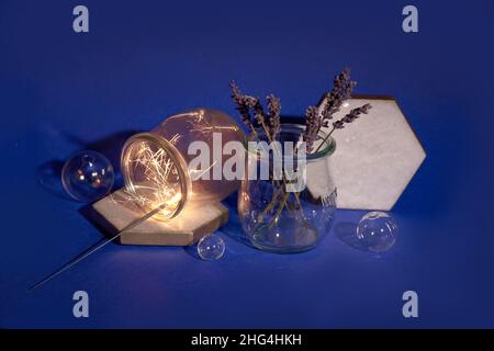 Dry lavender in glass, magic light with sparkles and orange glow. Dark blue, purple paper background. Light, long shadows. Simple minimal still life. Stock Photo