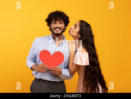 St. Valentine's day. Pretty indian woman kissing smiling man, holding red heart, yellow background Stock Photo